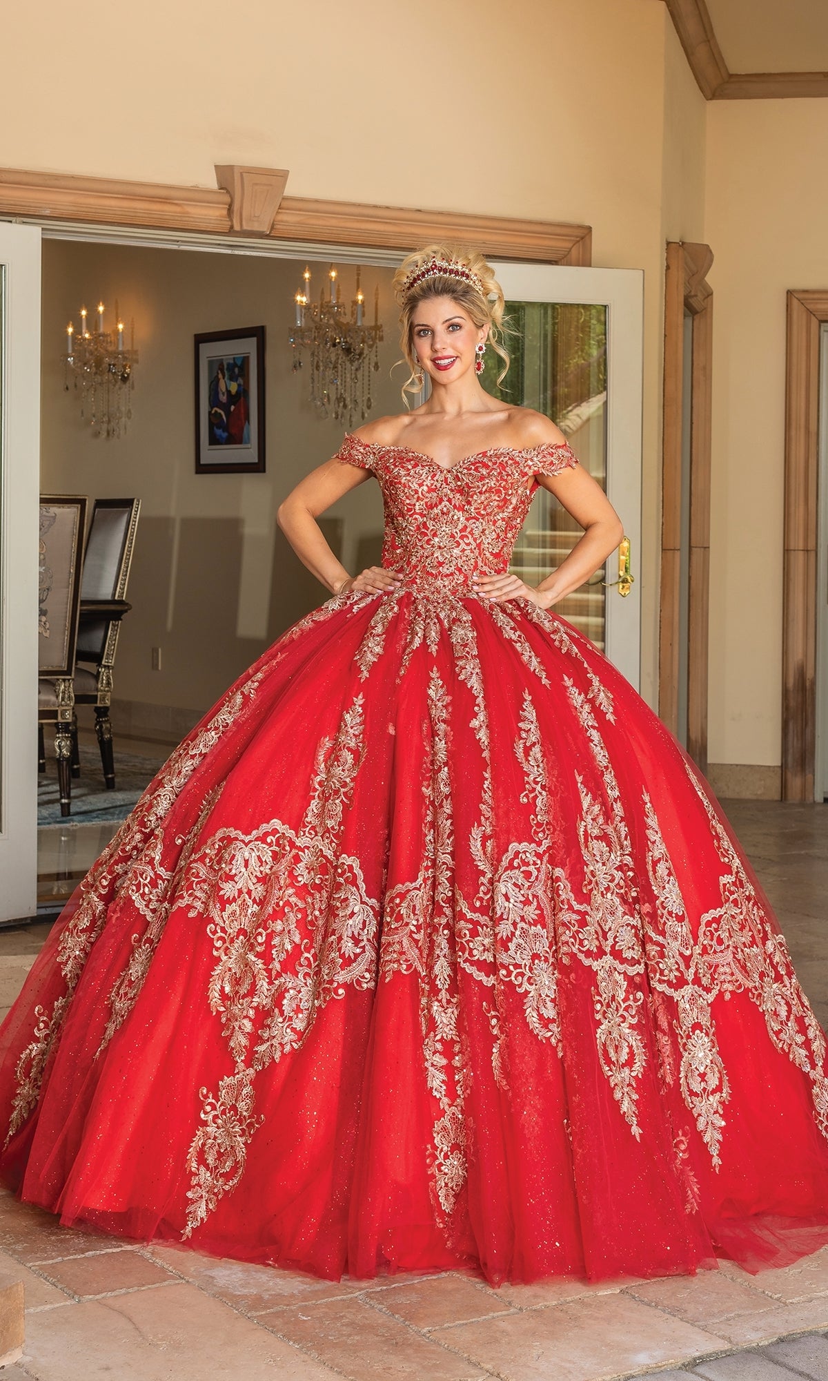A woman stands indoors wearing the Quinceanera Dress 1783 By Dancing Queen, featuring an off-the-shoulder design, gold lace embroidery, a sweetheart neckline, and a jeweled tiara.